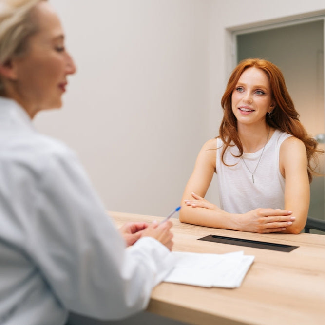 Rear view of smiling caucasian woman doctor listening young woman patient complaints, making notes in card clipboard or healthcare insurance form, at initial medical consultation appointment